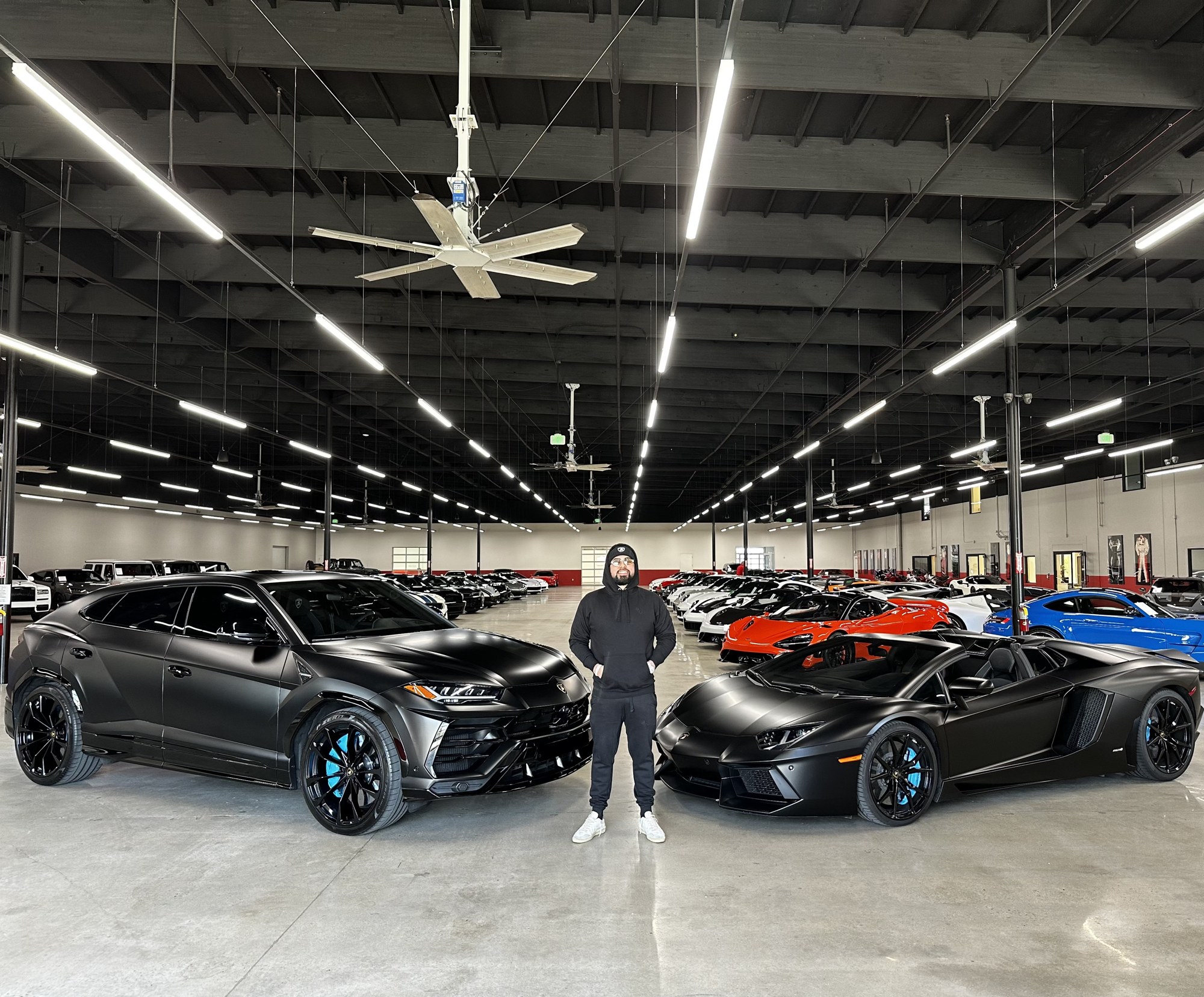 Evan Valdes standing between a black Lamborghini Urus and Aventador in a warehouse with a fleet behind him